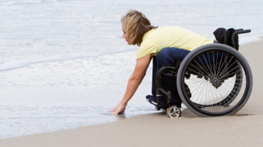 Person in a wheelchair on the beach, leaning down to touch the water